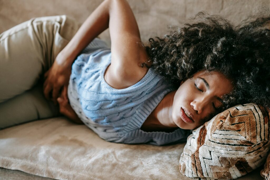 African American woman with an afro hairstyle experiencing severe stomach ulcer pain, lying down on a sofa with her eyes closed and hands on her abdomen