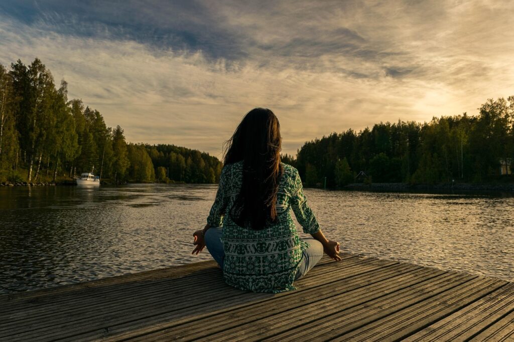 Just as the still water reflects the sky, a mind cleared of impurities through mindful control of thought reflects true peace. This is the essence of finding your inner freedom. A woman mastering mindful control of thought through meditation in lotus pose by a lake at sunset.