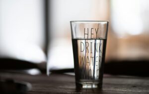 Your daily reminder: reaching for a glass of water is the easiest decision you'll make for your health. Each sip supports your energy, focus, and natural detoxification pathways. A close-up of a glass of clear water with the motivational text "Hey Drink Water More," illustrating the simple first step to unlocking the full benefits of drinking water for health and detox.