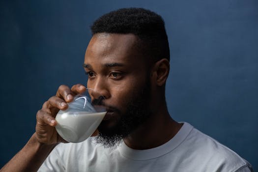 Mindful sipping is the key to how to digest milk better—slow drinking helps reduce bloating and makes milk easier on your stomach. A serious man in a white t-shirt drinks milk from a glass against a dark blue background, illustrating how to digest milk better by sipping slowly and mindfully to improve digestion and reduce discomfort.