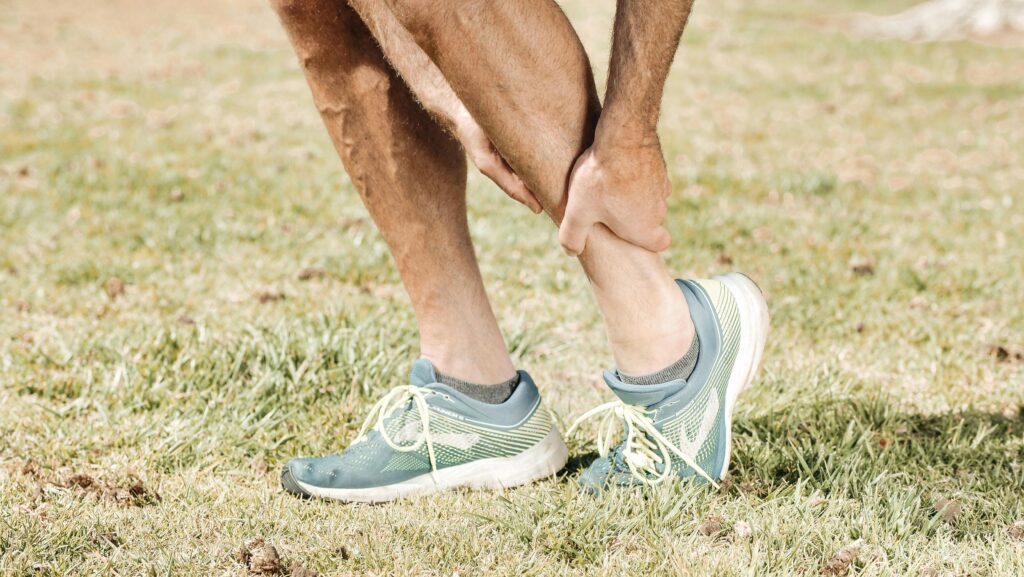 Man holding his calf in pain, wearing teal sneakers on green grass. Ankle pain from limited mobility—regular ankle circles prevent injury and improve flexibility.