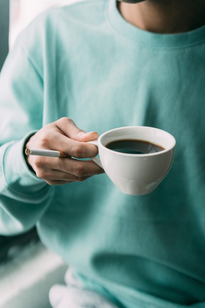 Adult casually enjoying a hot coffee and smoking indoors, wearing a turquoise sweater. An adult drinking coffee and smoking indoors, demonstrating habits to avoid when working to strengthen the heart naturally.