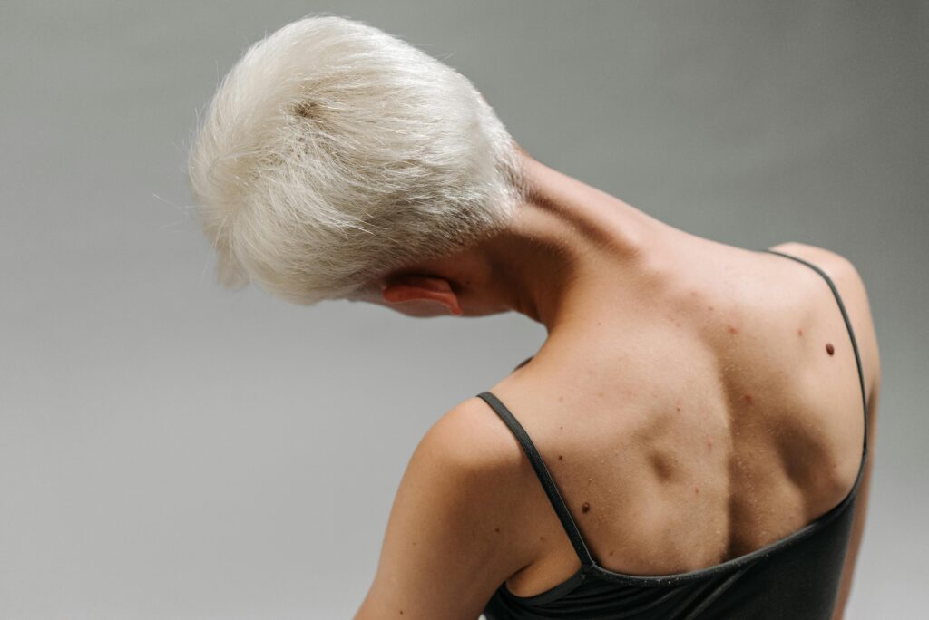 Back view of a woman with white hair bending her neck, showing body movement and detail on gray background. Neck circles mobility exercise demonstrated by senior woman on gray background.